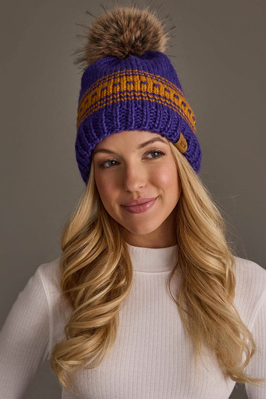 A woman with long blonde hair smiles softly against a plain gray background, wearing the Panache Apparel Collection's Purple & Gold Patterned Knit Pom Hat and a white ribbed long-sleeve top.
