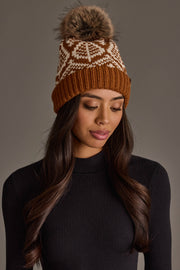A woman wears the Panache Apparel Collection Camel & Beige Patterned Knit Pom Hat, featuring a white geometric design and faux fur pom-pom, paired with a black ribbed long-sleeve top, as she looks down with a neutral expression against a plain background.