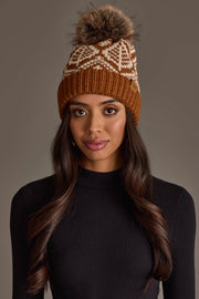 A woman with long, wavy brown hair wears the Panache Apparel Collection’s Camel & Beige Patterned Knit Pom Hat, featuring a white design and a fur pom-pom. She is dressed in a black, long-sleeve ribbed top and stands against a plain, neutral background.