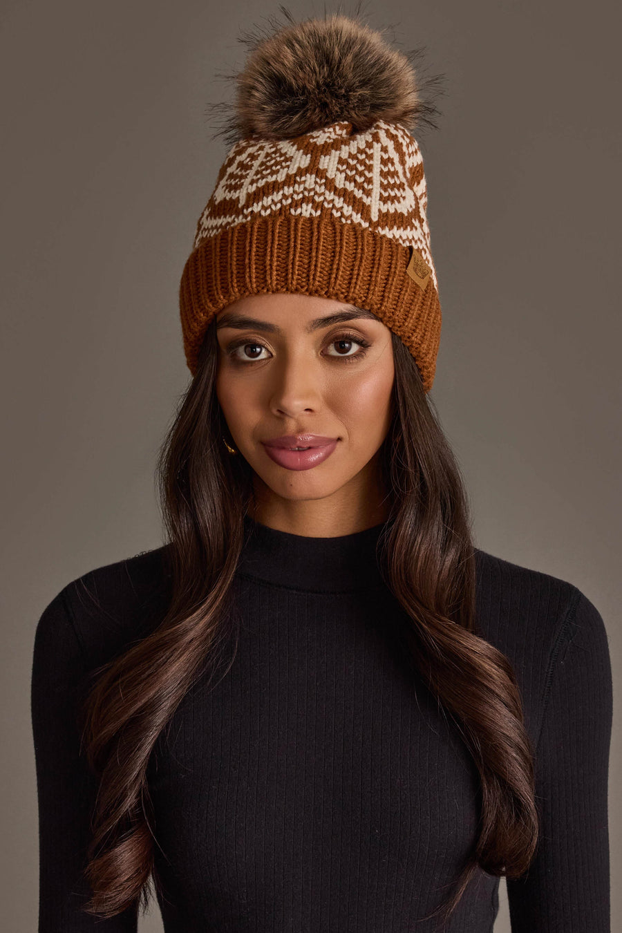 A woman with long, wavy brown hair wears the Panache Apparel Collection’s Camel & Beige Patterned Knit Pom Hat, featuring a white design and a fur pom-pom. She is dressed in a black, long-sleeve ribbed top and stands against a plain, neutral background.
