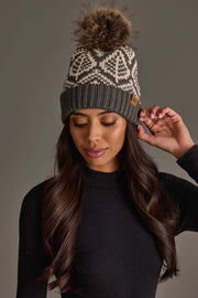 A woman with long dark hair, wearing a black sweater and the Panache Apparel Collection Gray & Beige Patterned Knit Pom Hat, featuring white designs and a faux fur pom-pom, looks down as she touches the side of her hat.