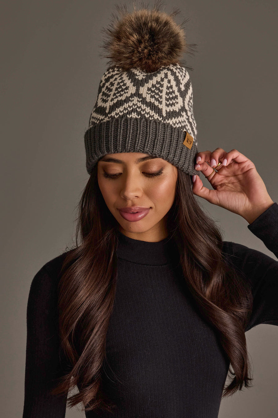 A woman with long dark hair, wearing a black sweater and the Panache Apparel Collection Gray & Beige Patterned Knit Pom Hat, featuring white designs and a faux fur pom-pom, looks down as she touches the side of her hat.