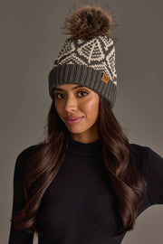 A woman with long dark hair is wearing a black sweater and the Gray & Beige Patterned Knit Pom Hat by Panache Apparel Collection, standing against a plain gray background.