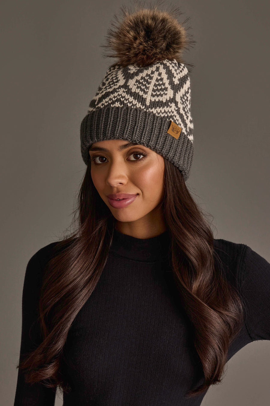 A woman with long dark hair is wearing a black sweater and the Gray & Beige Patterned Knit Pom Hat by Panache Apparel Collection, standing against a plain gray background.