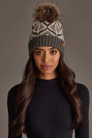 A woman with long dark hair wears a black knit sweater and the Panache Apparel Collection Gray & Beige Patterned Knit Pom Hat, featuring a geometric design and a large brown fur pom-pom, while standing against a plain gray background.