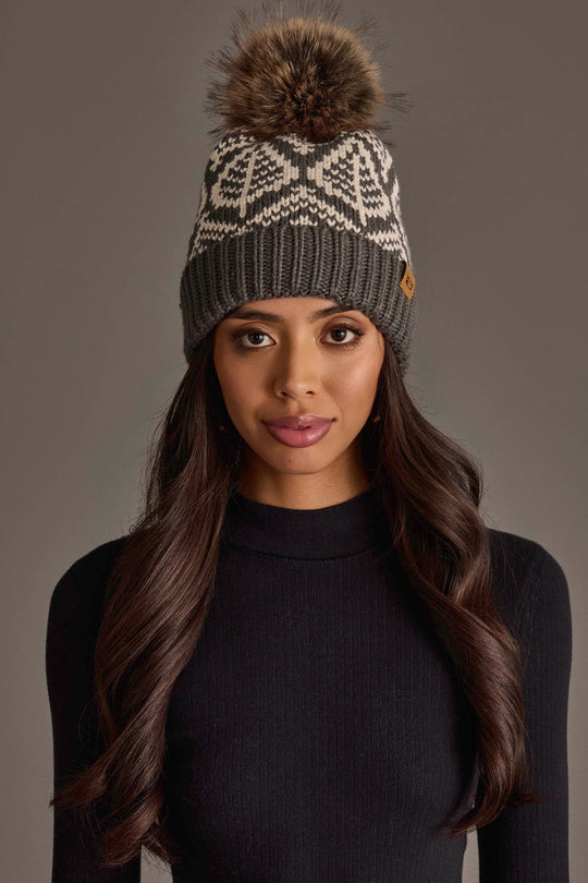 A woman with long dark hair wears a black knit sweater and the Panache Apparel Collection Gray & Beige Patterned Knit Pom Hat, featuring a geometric design and a large brown fur pom-pom, while standing against a plain gray background.