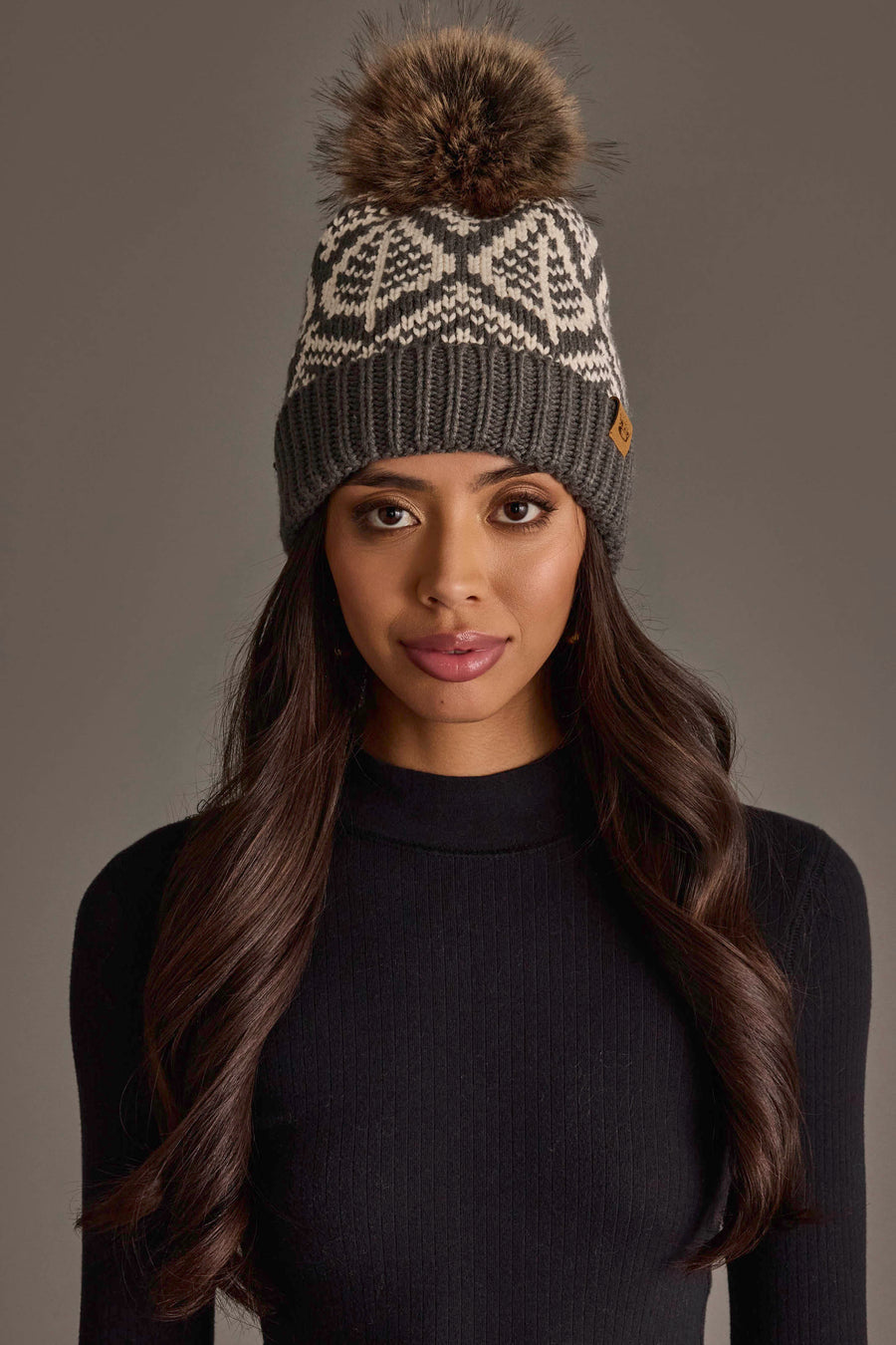 A woman with long dark hair wears a black knit sweater and the Panache Apparel Collection Gray & Beige Patterned Knit Pom Hat, featuring a geometric design and a large brown fur pom-pom, while standing against a plain gray background.