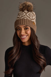 A woman with long dark hair, smiling in front of a neutral background, wears the Panache Apparel Collection’s Taupe & Beige Patterned Knit Pom Hat along with a black top.