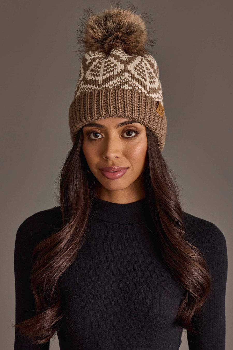 A woman with long, wavy dark hair wears the Taupe & Beige Patterned Knit Pom Hat by Panache Apparel Collection, featuring a white pattern and faux fur pom-pom, paired with a black sweater and standing against a neutral gray background.