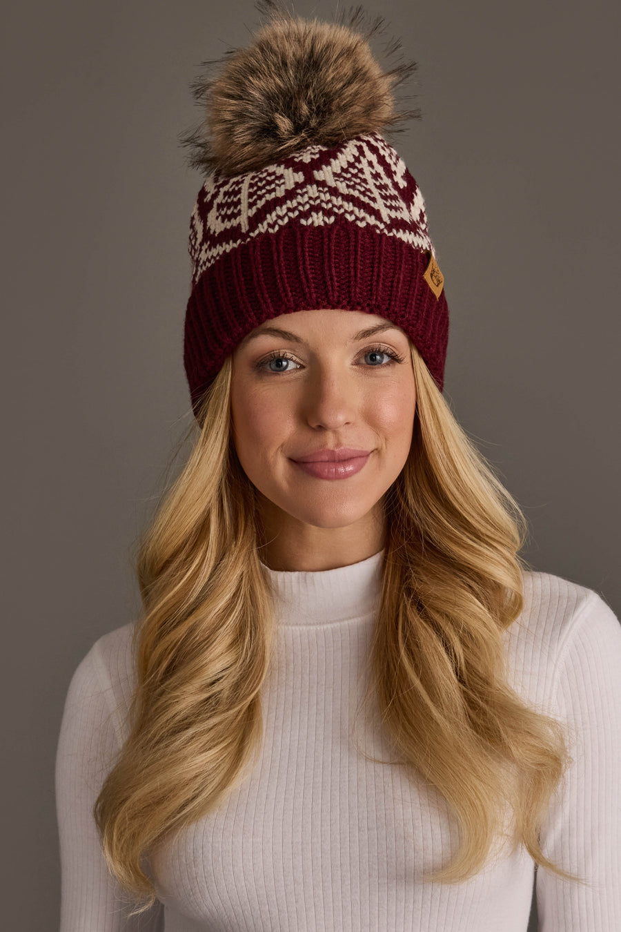 A woman with long blonde hair poses against a plain gray background, wearing the Panache Apparel Collection’s Wine & Beige Patterned Knit Pom Hat with a furry pom-pom and a white ribbed turtleneck sweater.