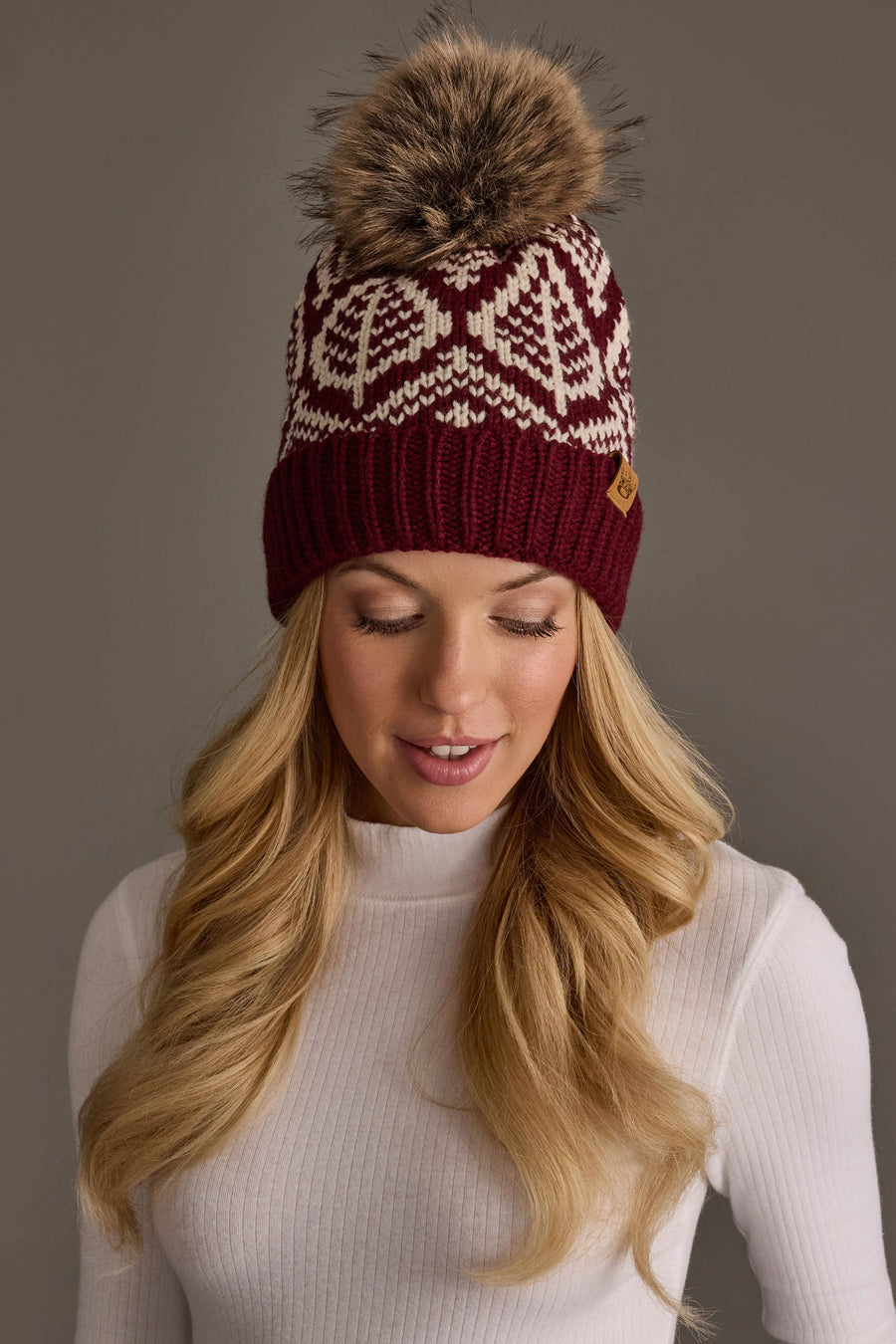 A woman with long blonde hair wears the Panache Apparel Collection Wine & Beige Patterned Knit Pom Hat, featuring a white geometric design and a large faux fur pom-pom, paired with a white ribbed turtleneck sweater. She is looking down against a plain background.