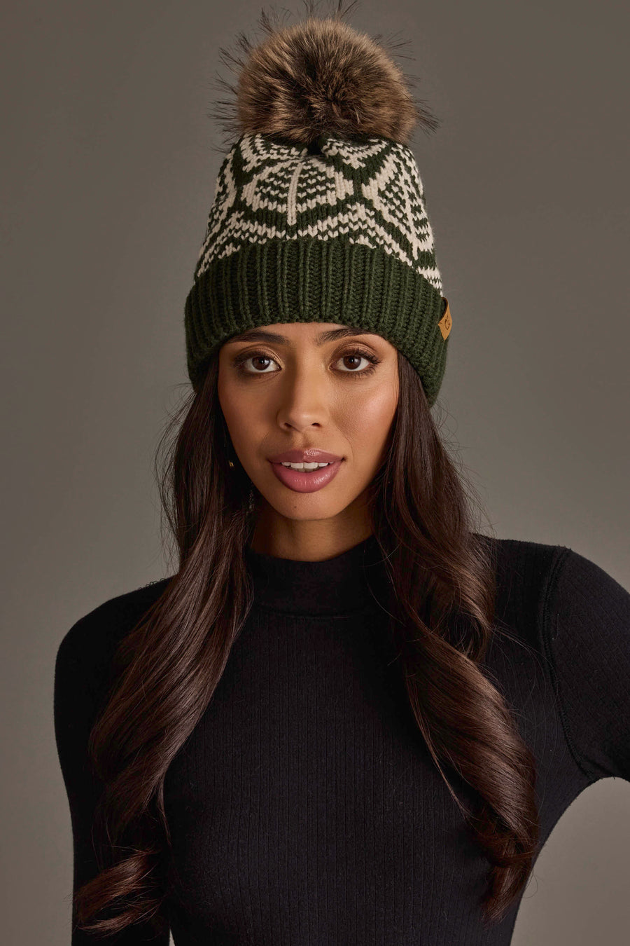 A woman with long brown hair wears the Panache Apparel Collection Olive & Beige Patterned Knit Pom Hat, featuring a fur pom-pom, paired with a black ribbed sweater, as she poses against a plain grey background.