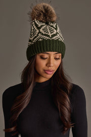 A woman with long brown hair wears the Olive & Beige Patterned Knit Pom Hat from Panache Apparel Collection, featuring a white geometric design and faux fur pom-pom. She pairs it with a black ribbed sweater and looks down with a neutral expression against a plain background.