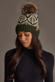 A woman wearing the Panache Apparel Collection Olive & Beige Patterned Knit Pom Hat, featuring a white geometric design and a faux fur pom-pom, looks down. She has long, wavy dark hair and is dressed in a black ribbed top against a neutral backdrop.