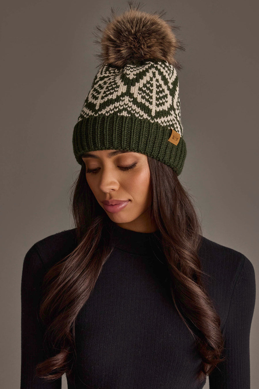 A woman wearing the Panache Apparel Collection Olive & Beige Patterned Knit Pom Hat, featuring a white geometric design and a faux fur pom-pom, looks down. She has long, wavy dark hair and is dressed in a black ribbed top against a neutral backdrop.