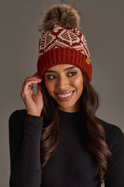 A smiling woman with long dark hair wears the Panache Apparel Collection’s Rust & Beige Patterned Knit Pom Hat, featuring a furry pom-pom, along with a black ribbed turtleneck shirt, against a plain gray background.