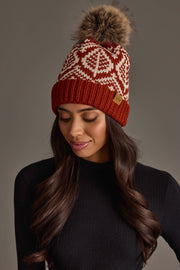 A woman from the Panache Apparel Collection wears the Rust & Beige Patterned Knit Pom Hat, featuring a stylish snowflake design and a faux fur pom-pom. She smiles as she looks down, paired with a black ribbed sweater against a neutral background.