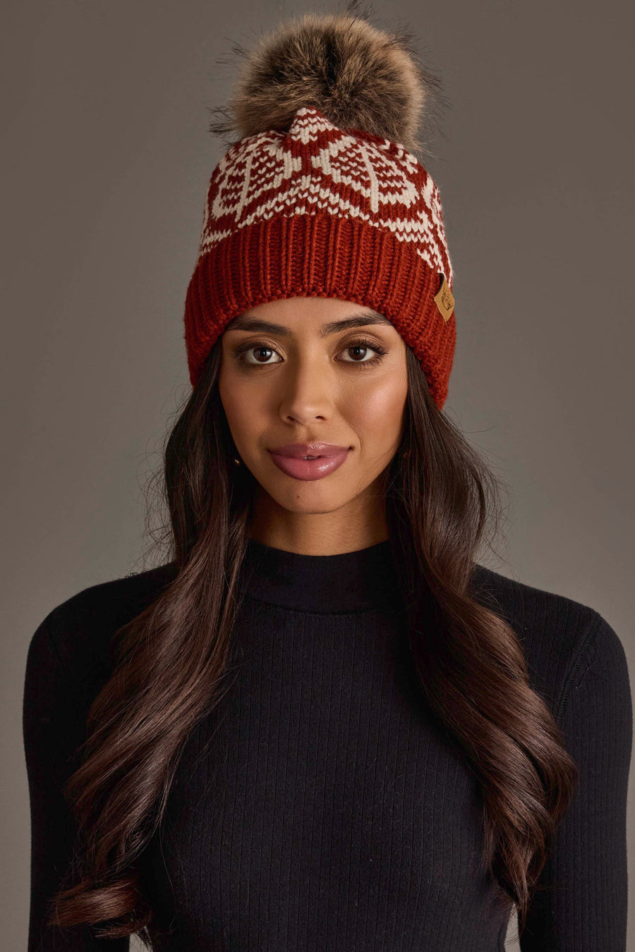 A woman wearing the Panache Apparel Collection’s Rust & Beige Patterned Knit Pom Hat with a faux fur pom-pom, paired with a black long-sleeve top, stands against a plain gray background.