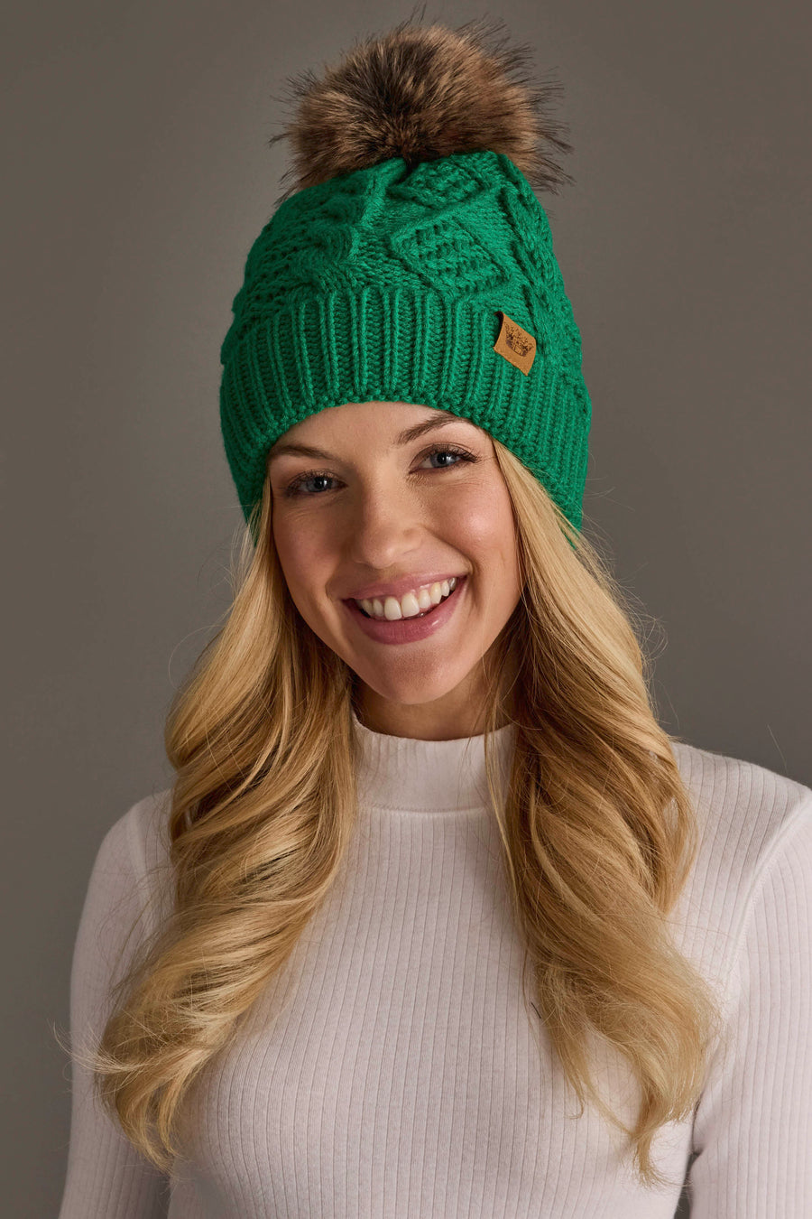 A smiling woman with long blonde hair models the Panache Apparel Collection Bright Classic Green Cable Knit Pom Hat, featuring a brown pom-pom, paired with a white ribbed long-sleeve top and posed against a plain gray background.