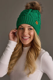 A smiling woman with long blonde hair wears the Panache Apparel Collection’s Bright Classic Green Cable Knit Pom Hat, featuring a brown pom-pom and a small leather patch, along with a white ribbed long-sleeve top, as she poses against a plain gray background.