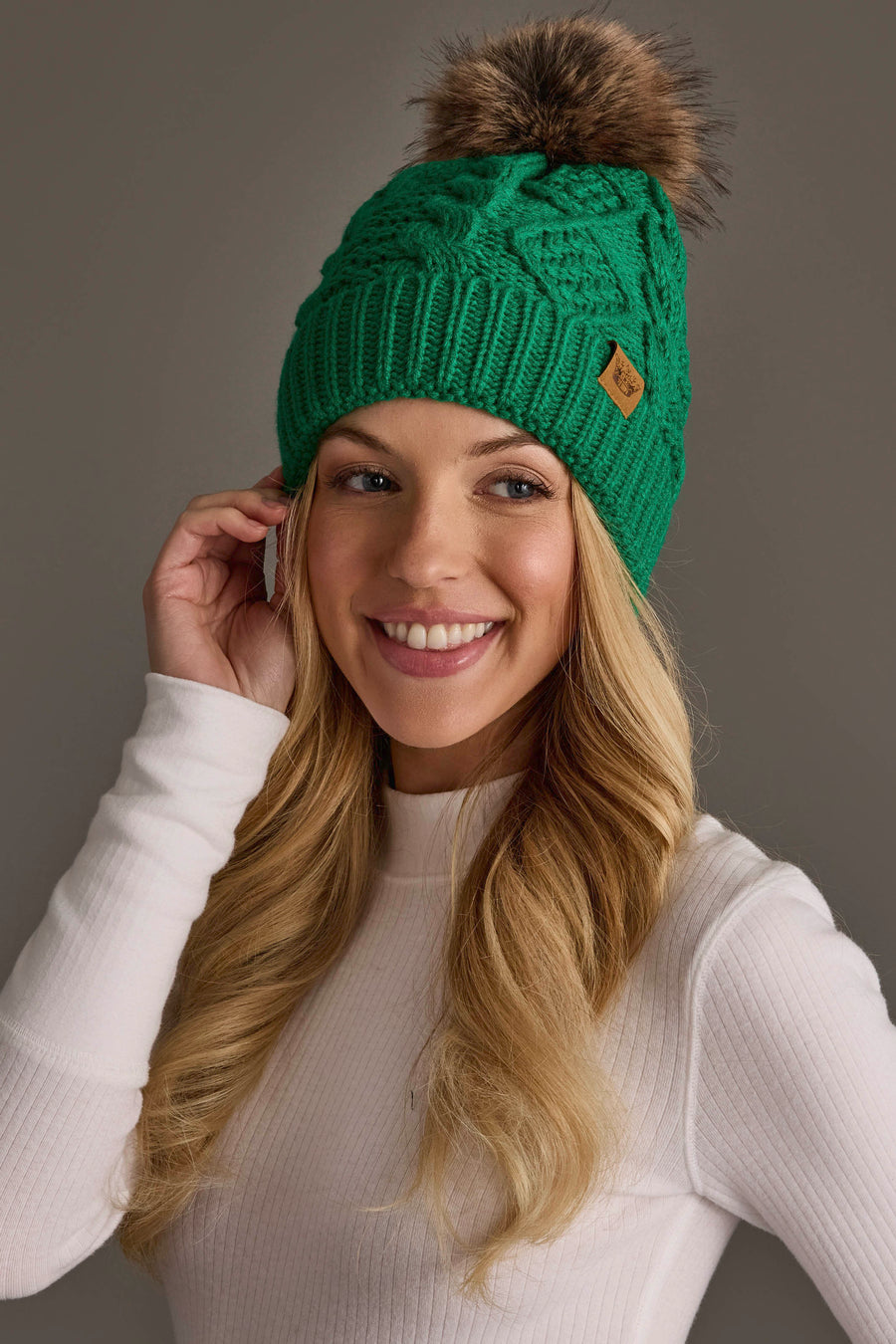 A smiling woman with long blonde hair wears the Panache Apparel Collection’s Bright Classic Green Cable Knit Pom Hat, featuring a brown pom-pom and a small leather patch, along with a white ribbed long-sleeve top, as she poses against a plain gray background.