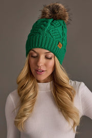 A woman with long blonde hair is wearing the Panache Apparel Collection Bright Classic Green Cable Knit Pom Hat, featuring a brown fur pom-pom, along with a white ribbed sweater. She looks down with a neutral expression against a plain gray background.
