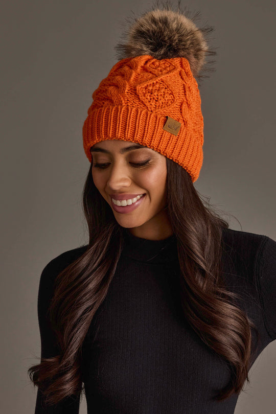 A woman with long brown hair is smiling and looking down while wearing the Panache Apparel Collection Orange Cable Knit Pom Hat and a black ribbed sweater.