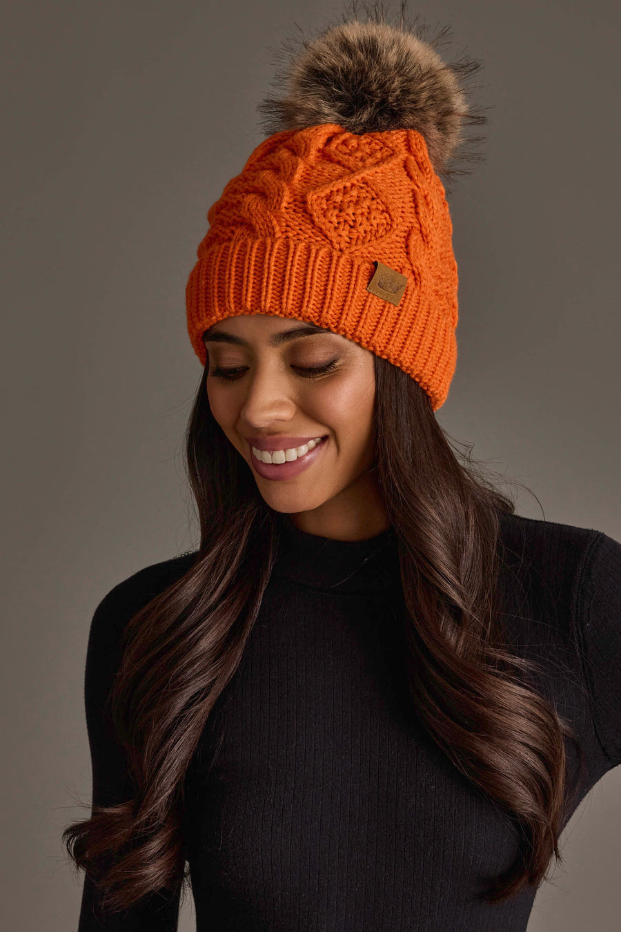 A woman with long brown hair is smiling and looking down while wearing the Panache Apparel Collection Orange Cable Knit Pom Hat and a black ribbed sweater.