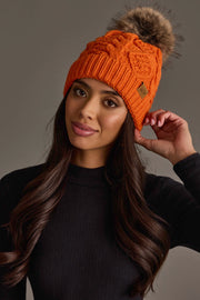 A woman with long dark hair, dressed in a black sweater, looks at the camera and touches her Panache Apparel Collection Orange Cable Knit Pom Hat, featuring a fur pom-pom, against a plain gray background.