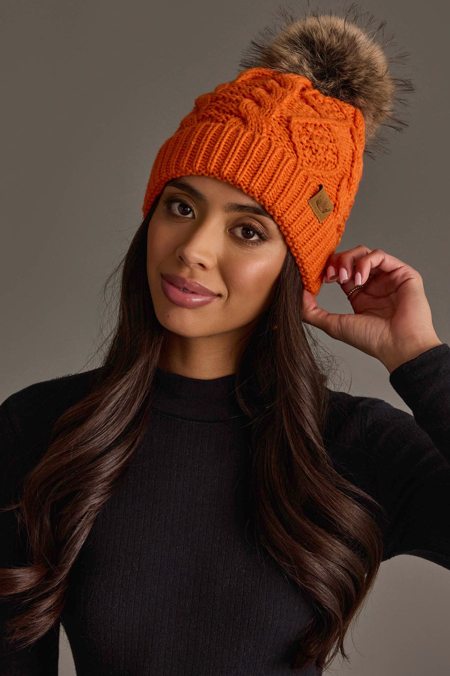 A woman with long dark hair, dressed in a black sweater, looks at the camera and touches her Panache Apparel Collection Orange Cable Knit Pom Hat, featuring a fur pom-pom, against a plain gray background.