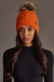A woman with long dark hair poses against a plain brown background wearing the Panache Apparel Collection Orange Cable Knit Pom Hat with a large faux fur pom-pom and a black ribbed sweater.