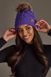 A smiling woman with long brown hair wears the Panache Apparel Collection Purple Cable Knit Pom Hat, featuring a faux fur pom-pom, styled with a black turtleneck and gold rings, as she poses with her hands near the hat against a neutral background.
