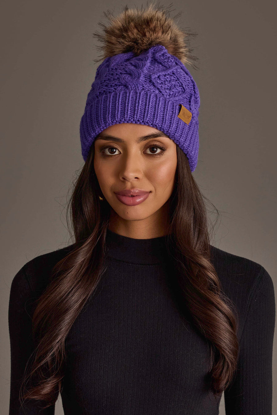 A woman with long brown hair wearing the Panache Apparel Collection Purple Cable Knit Pom Hat and a black ribbed turtleneck sweater, posing against a neutral background.