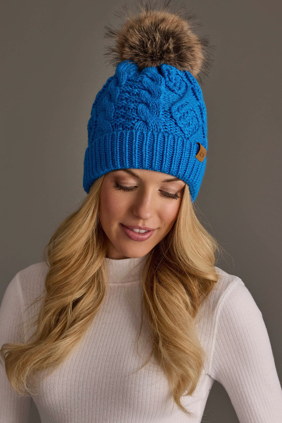 A woman with long blonde hair is seen looking down and smiling softly against a neutral background, wearing the Panache Apparel Collection’s Blue Cable Knit Pom Hat and a white ribbed turtleneck.