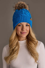 A woman with long blonde hair is smiling in front of a plain, neutral background while wearing the Panache Apparel Collection Blue Cable Knit Pom Hat and a white ribbed long-sleeve shirt.