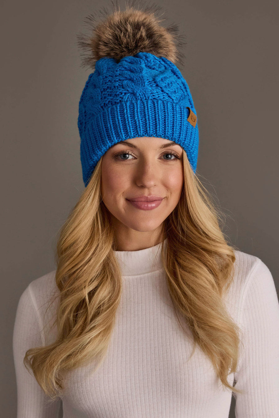 A woman with long blonde hair is smiling in front of a plain, neutral background while wearing the Panache Apparel Collection Blue Cable Knit Pom Hat and a white ribbed long-sleeve shirt.