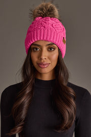A woman with long brown hair models the Bright Pink Cable Knit Pom Hat by Panache Apparel Collection, featuring a brown pom-pom. She pairs it with a black ribbed sweater and poses against a plain gray background.