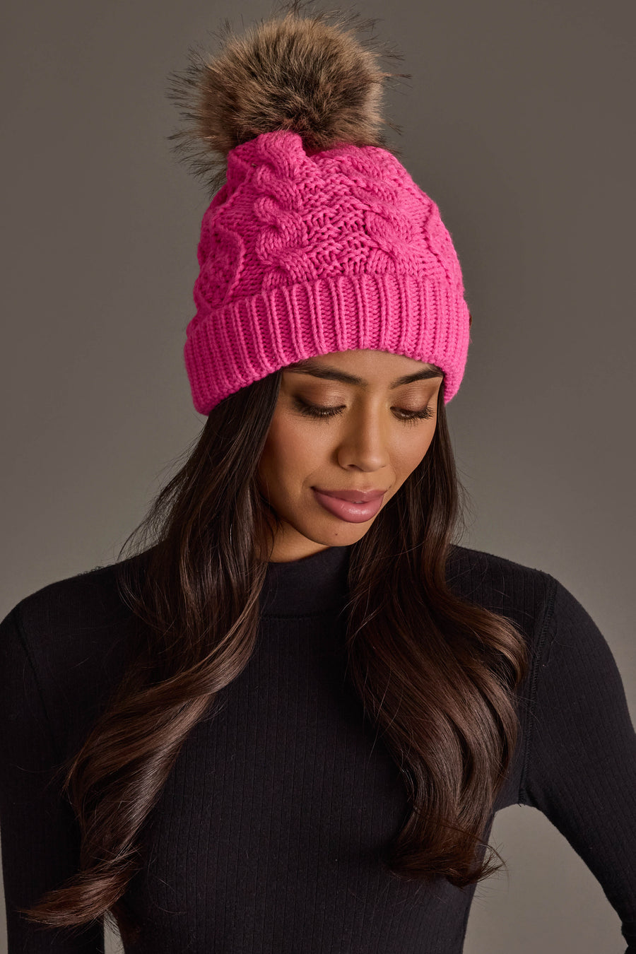 A woman with long brown hair is seen looking down with a neutral expression against a gray background, wearing the Panache Apparel Collection’s Bright Pink Cable Knit Pom Hat featuring a brown faux fur pom-pom, paired with a black long-sleeve top.