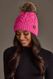 A woman with long brown hair is smiling and looking down, dressed in a black sweater and wearing the Panache Apparel Collection Bright Pink Cable Knit Pom Hat, featuring a fur pom-pom, against a plain background.