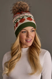 A woman with long blonde hair wears a white ribbed turtleneck and the Panache Apparel Collection Olive, Cream, Rust & Pink Patterned Pom Hat, featuring a brown faux fur pom-pom; she smiles and looks slightly to the side.