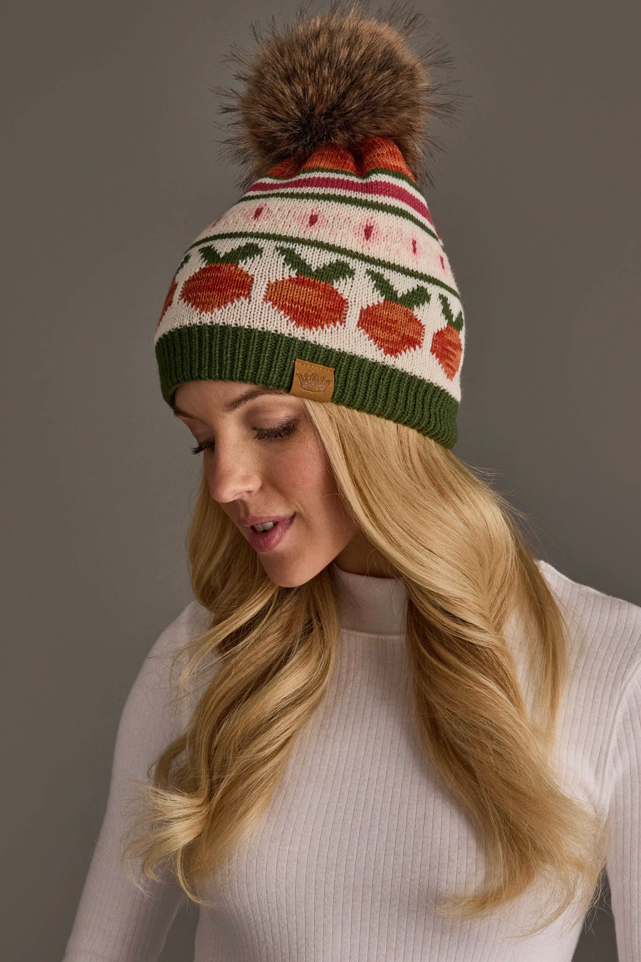 A woman with long blonde hair wears a white ribbed sweater and the Panache Apparel Collection Olive, Cream, Rust & Pink Patterned Pom Hat, a knit beanie with colorful patterns and a large brown faux fur pom-pom, posing against a neutral background, looking downward with a gentle smile.