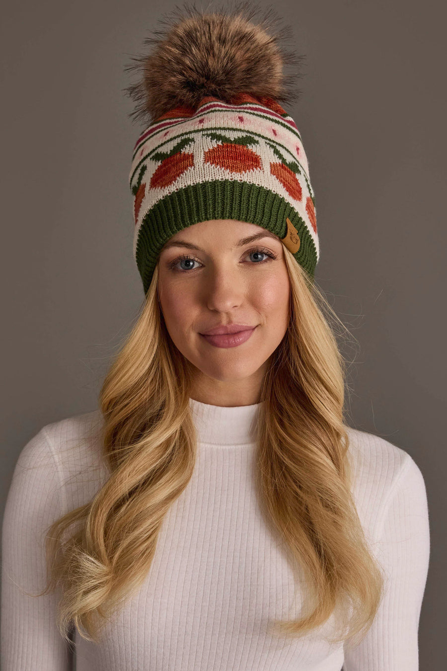 A woman with long blonde hair wears a white ribbed sweater and the Panache Apparel Collection’s Olive, Cream, Rust & Pink Patterned Pom Hat, a colorful knit beanie adorned with a pom-pom,while standing against a plain gray background.