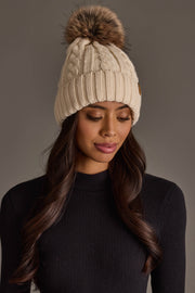 A woman with long wavy hair wears the Panache Apparel Collection’s Cream Cable Knit Design Pom Hat and a black ribbed sweater. She is looking down with a neutral expression against a plain gray background.