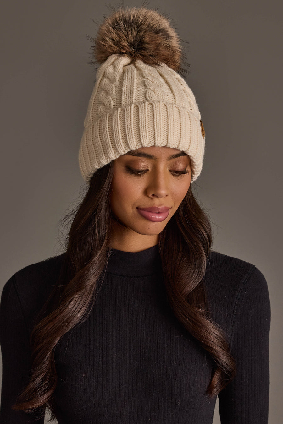 A woman with long wavy hair wears the Panache Apparel Collection’s Cream Cable Knit Design Pom Hat and a black ribbed sweater. She is looking down with a neutral expression against a plain gray background.