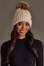 A woman with long dark hair smiles against a plain gray background, wearing the Panache Apparel Collection Cream Cable Knit Design Pom Hat and a black ribbed turtleneck.