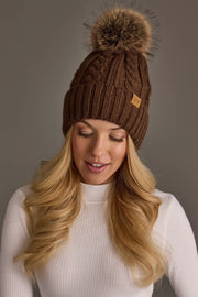 A woman with long blonde hair wears the Panache Apparel Collection’s Brown Cable Knit Design Pom Hat, featuring a large faux fur pom-pom, and a white ribbed turtleneck sweater. She smiles softly while looking down against a plain gray background.