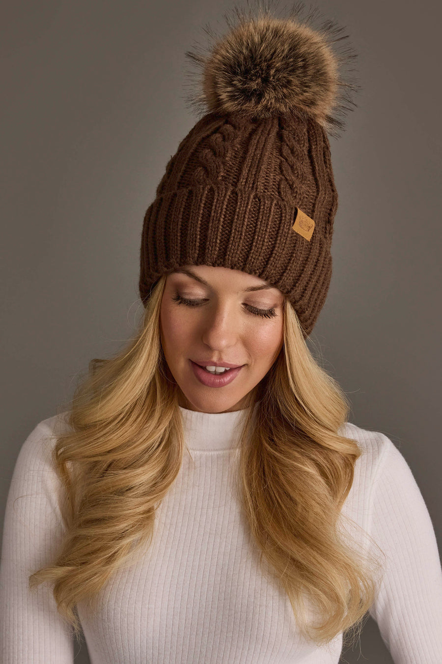 A woman with long blonde hair wears the Panache Apparel Collection’s Brown Cable Knit Design Pom Hat, featuring a large faux fur pom-pom, and a white ribbed turtleneck sweater. She smiles softly while looking down against a plain gray background.