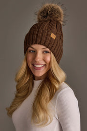 A woman with long blonde hair, wearing the Panache Apparel Collection’s Brown Cable Knit Design Pom Hat, smiles while looking slightly to the side. She is dressed in a white ribbed long-sleeve top against a neutral background.