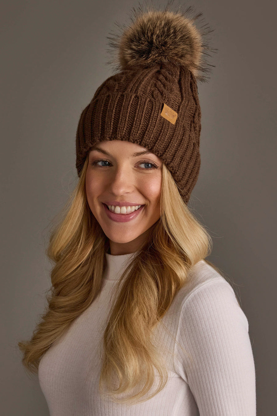 A woman with long blonde hair, wearing the Panache Apparel Collection’s Brown Cable Knit Design Pom Hat, smiles while looking slightly to the side. She is dressed in a white ribbed long-sleeve top against a neutral background.