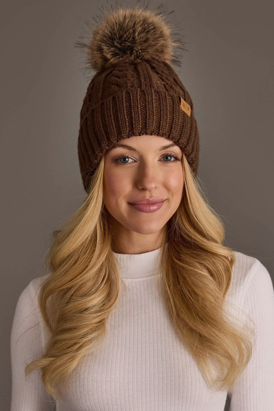 A woman with long blonde hair smiles softly against a neutral background, wearing the Panache Apparel Collection Brown Cable Knit Design Pom Hat and a white ribbed long-sleeve top.